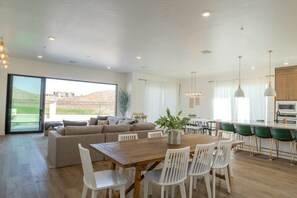 Uma sala ampla com um sofá grande, mesa de jantar e cadeiras. Há uma cozinha com um balcão e vista para o exterior.