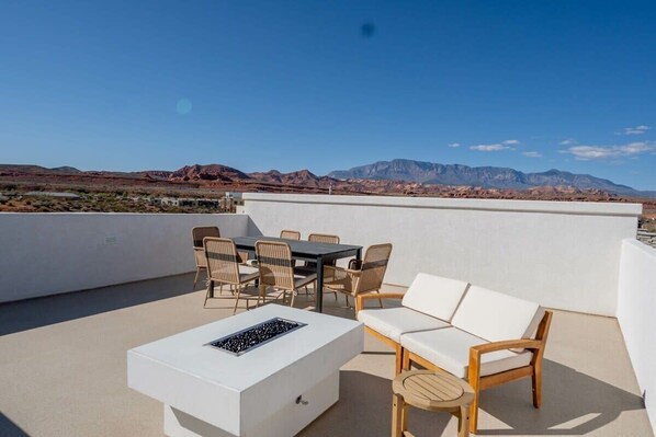 A rooftop terrace with a white table, chairs, and a sofa, overlooking a mountainous landscape.