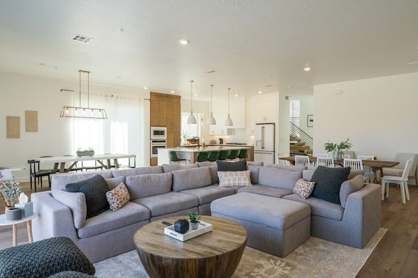 A spacious living room with a large grey sectional sofa, a wooden coffee table, and a dining area with a white table and chairs.