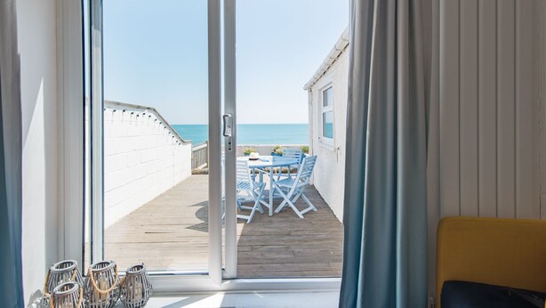 A room with a sliding glass door leading to a wooden deck with a table and chairs, overlooking a beach. - Wave Cottage -  seaside cottage right on the beach by Bloom Stays (Sandgate)