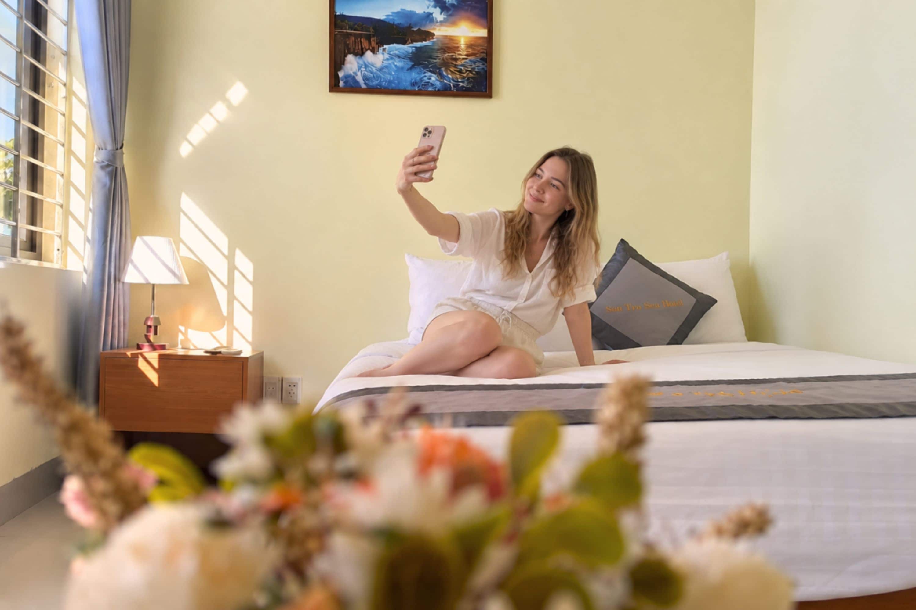 A person sitting on a bed in a room with a nightstand, a lamp, and a painting on the wall.