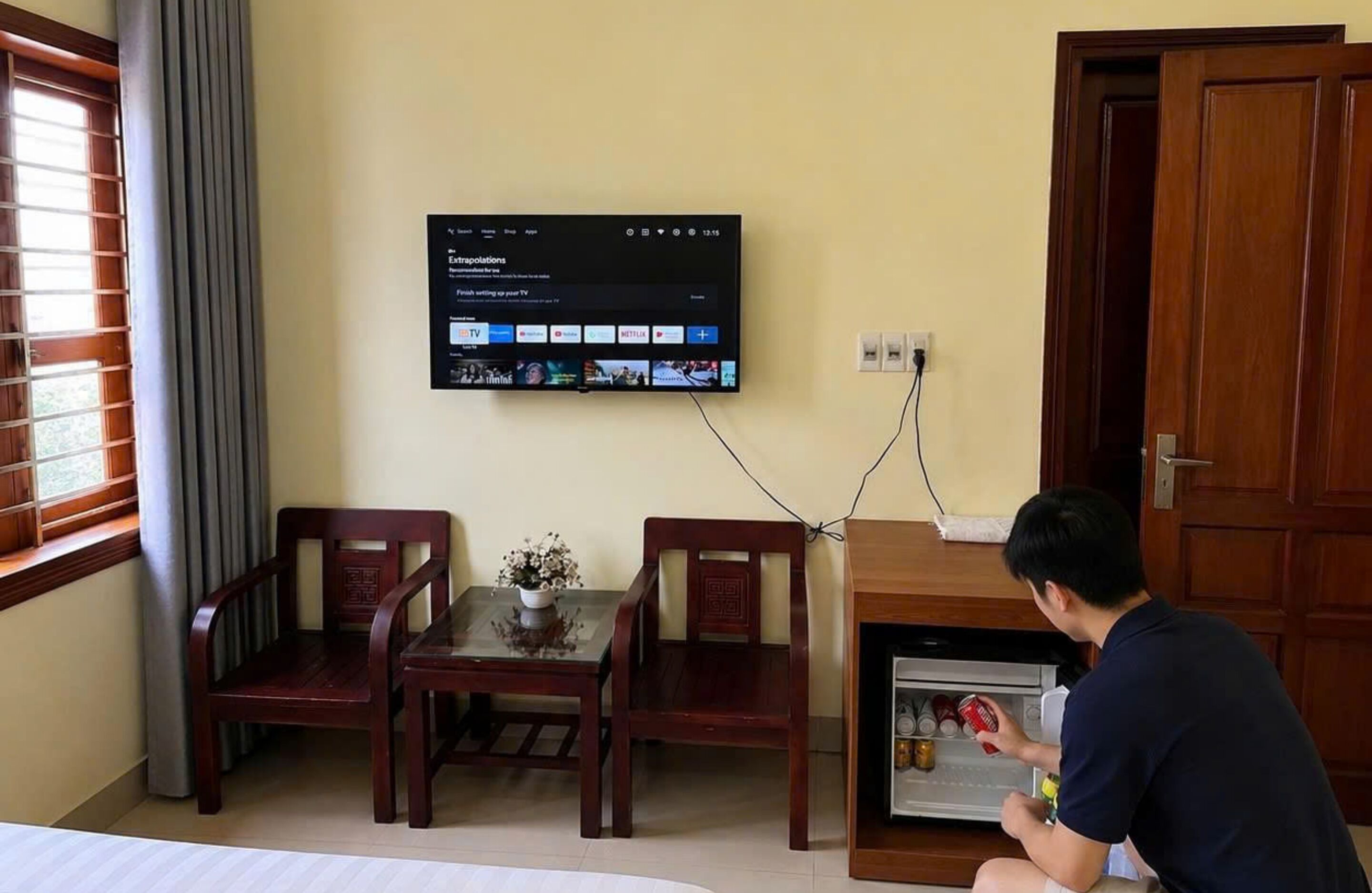 Una camera d'albergo con una televisione a parete che mostra l'interfaccia di un servizio di streaming, un tavolino con un vaso, due sedie di legno e una persona seduta sul pavimento vicino a un frigorifero.