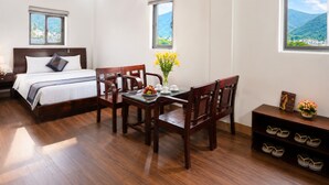 A hotel room with a bed, a dining table with chairs, a small table with a vase of flowers, a fan, and a window with a mountain view. - SONTRA SEA HOTEL (Da Nang)