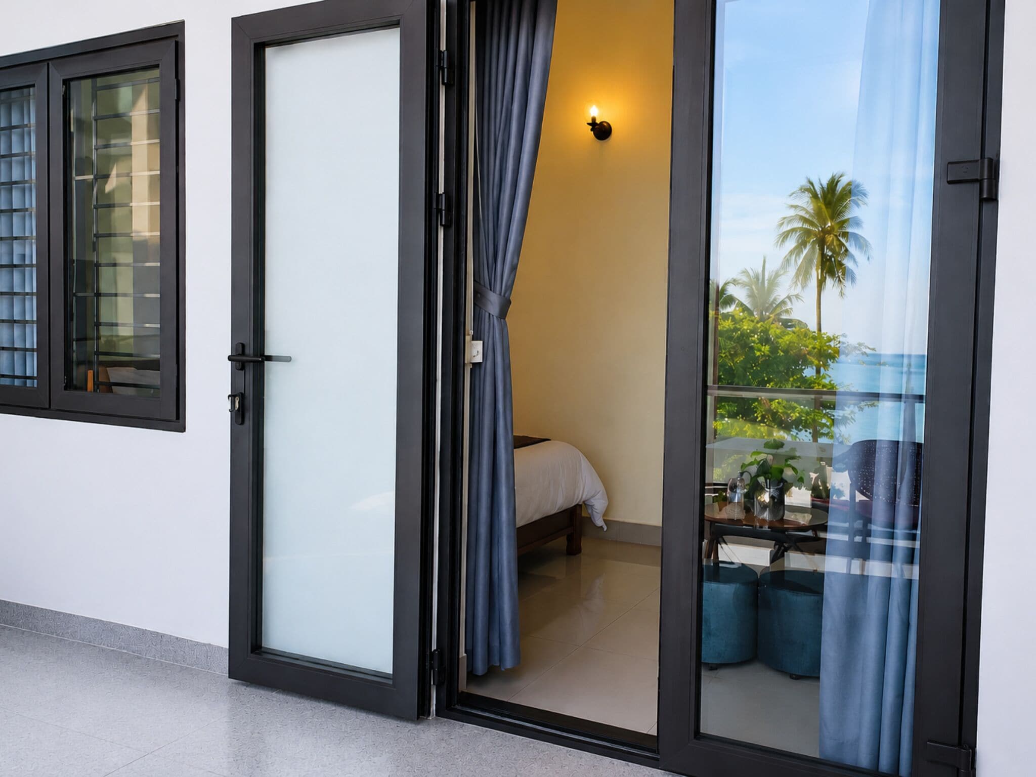 A room with a bed, a small table, a chair, and a balcony with a view of palm trees and the sea.