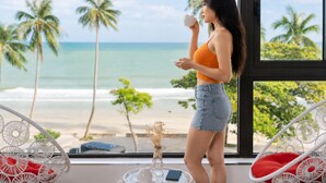 A room with a large window overlooking a beach, a wicker chair with red cushions, and a small table with a vase and a cup. - SONTRA SEA HOTEL (Da Nang)
