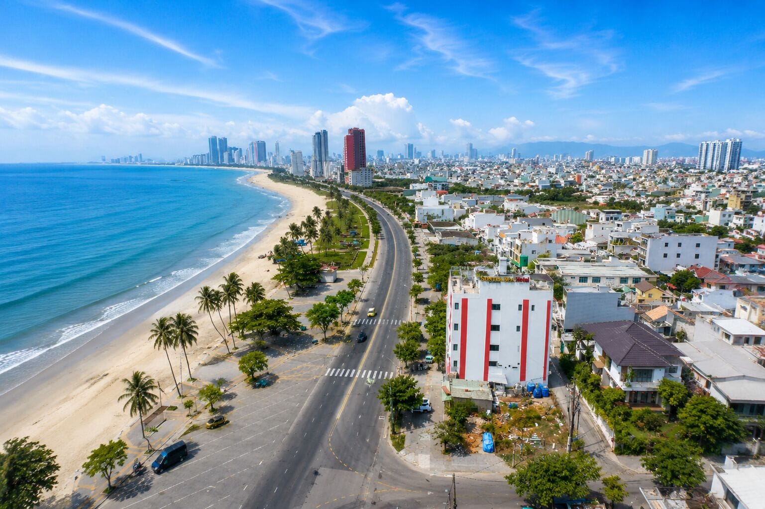 A coastal cityscape with a beach, a road curving along the shore, and a hotel with a red and white facade.