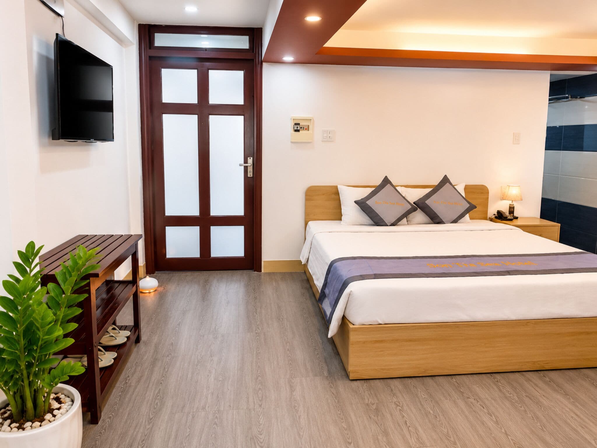 A hotel room with a bed, bedside table, lamp, potted plant, and a TV mounted on the wall.