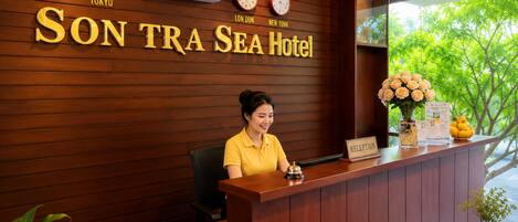 A reception desk at SON TRA SEA Hotel with clocks displaying different time zones, a receptionist, and a window with a view of trees.