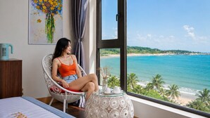 A room with a large window, a bed, a chair, a table with a vase, and a painting on the wall. - SONTRA SEA HOTEL (Da Nang)