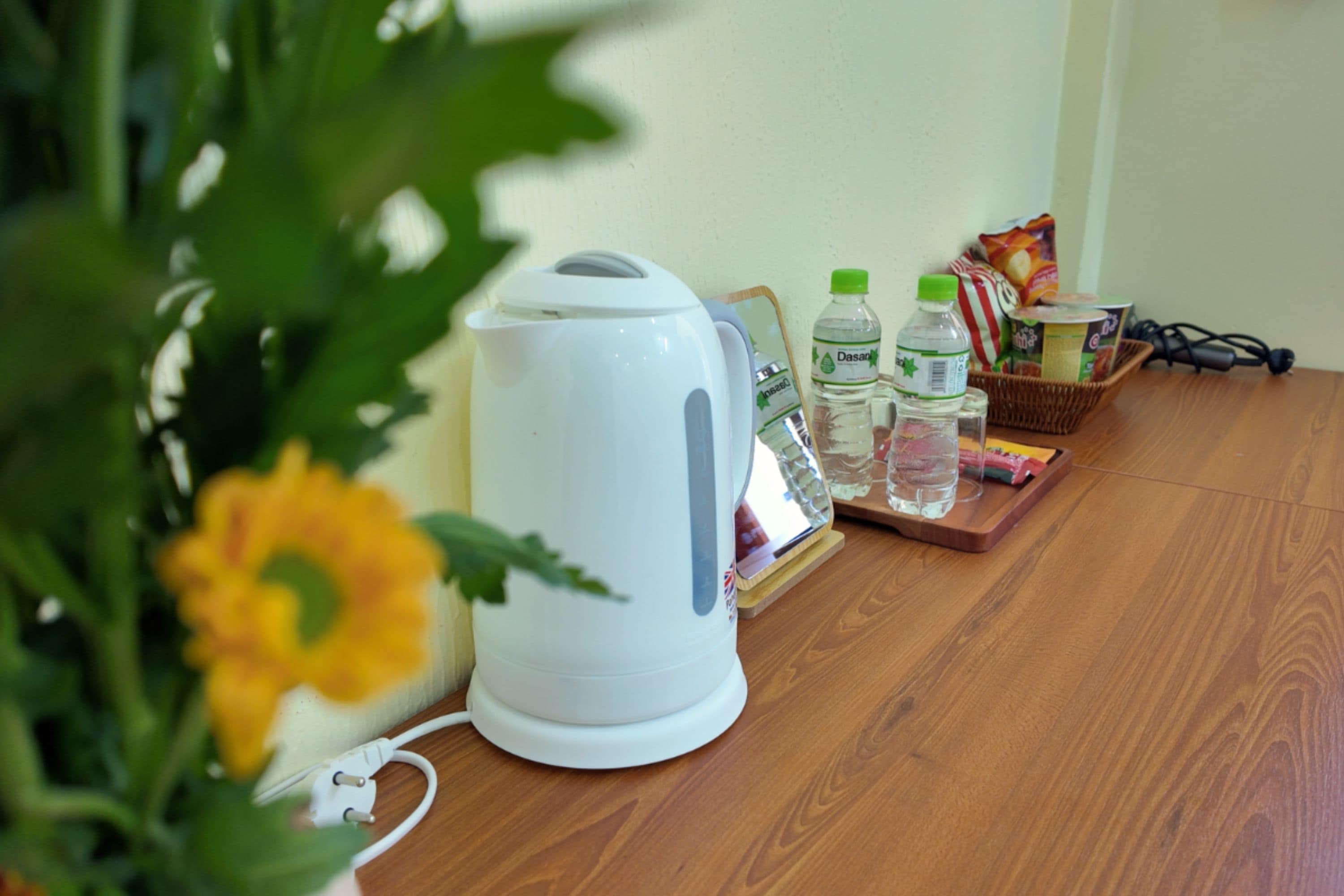 Une bouilloire électrique blanche, deux bouteilles d’eau Dasani, un panier rempli d’en-cas et un câble d’alimentation sur une table en bois.