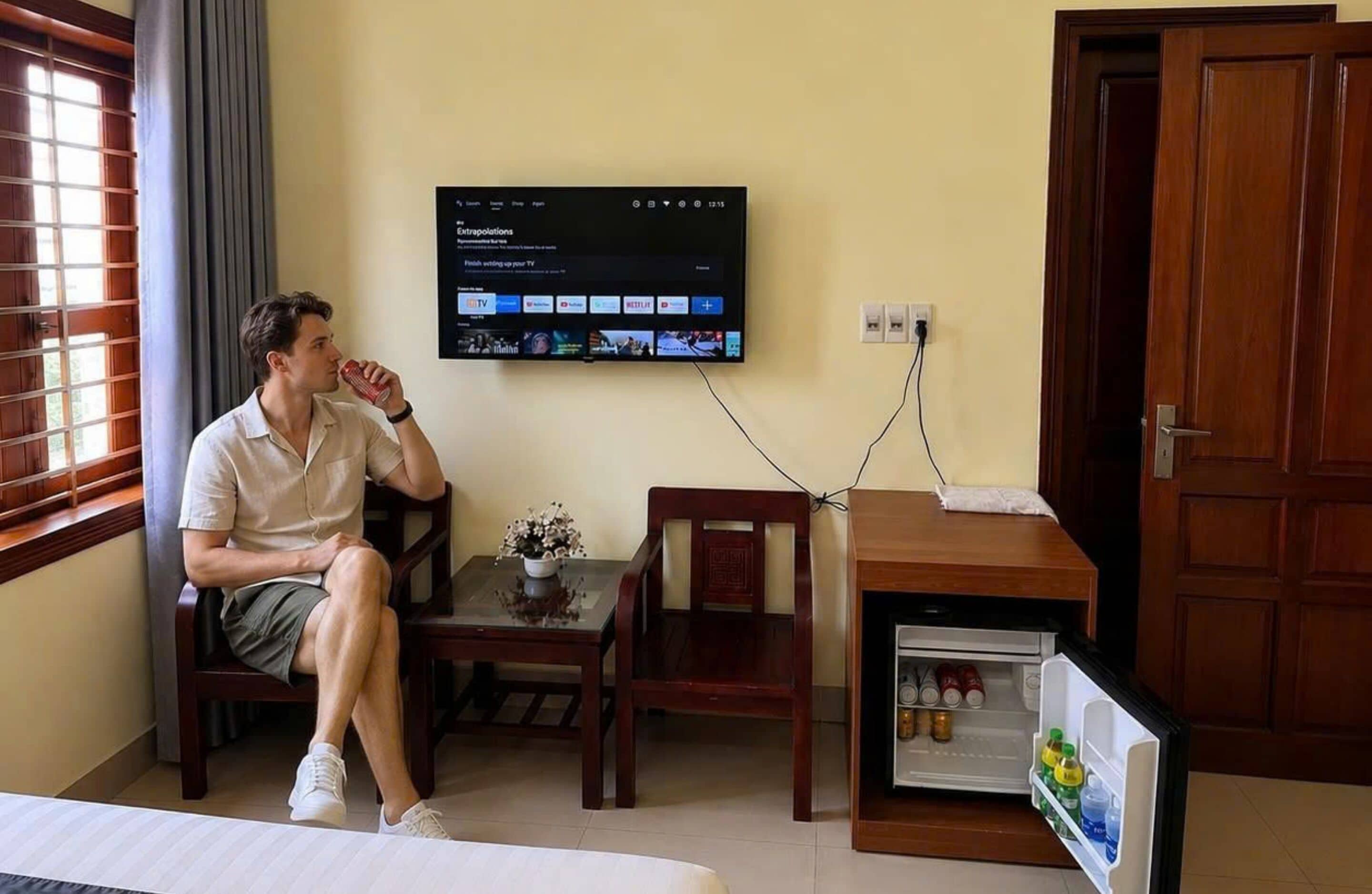 Una camera d'albergo con un letto, una TV a muro, un tavolino con un vaso, una porta in legno e un frigorifero.