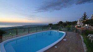 A swimming pool with a view of the sea, surrounded by a wooden deck and glass railing.