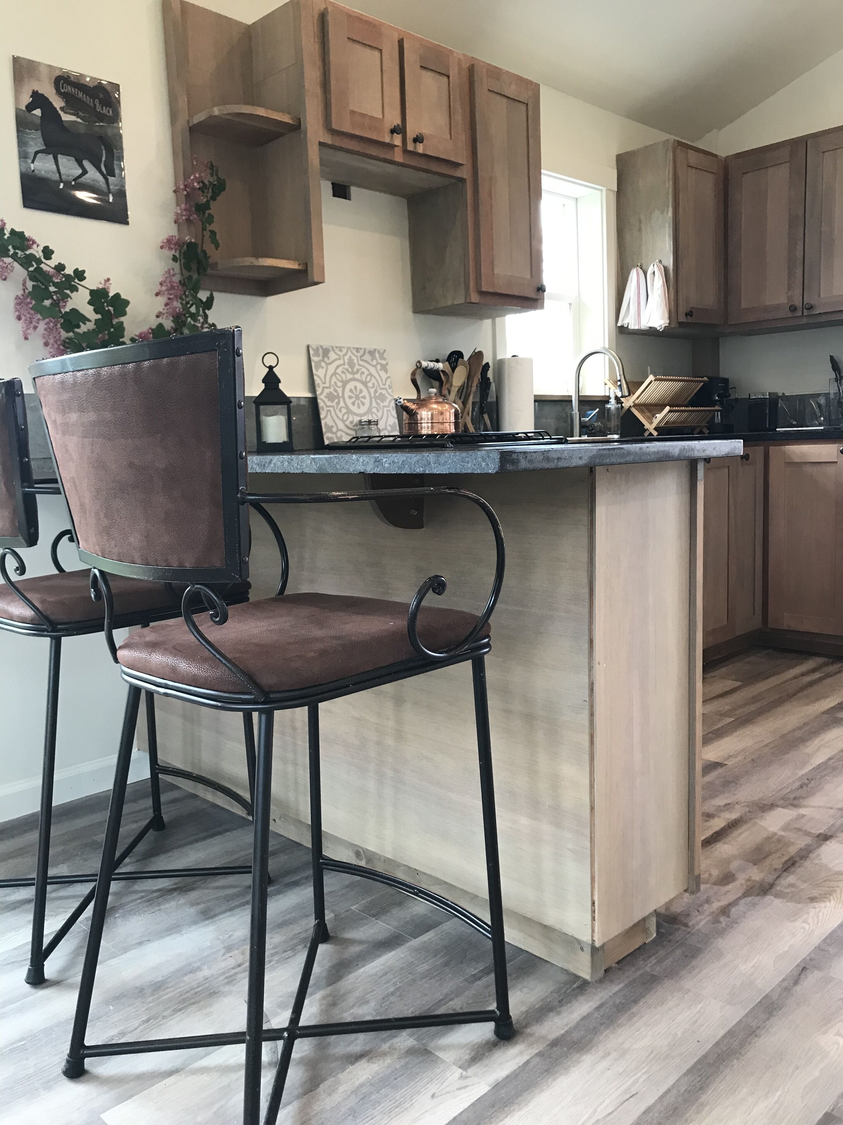 Een keuken met houten kastjes, een barstoel, een koperen pan en een hangplant.