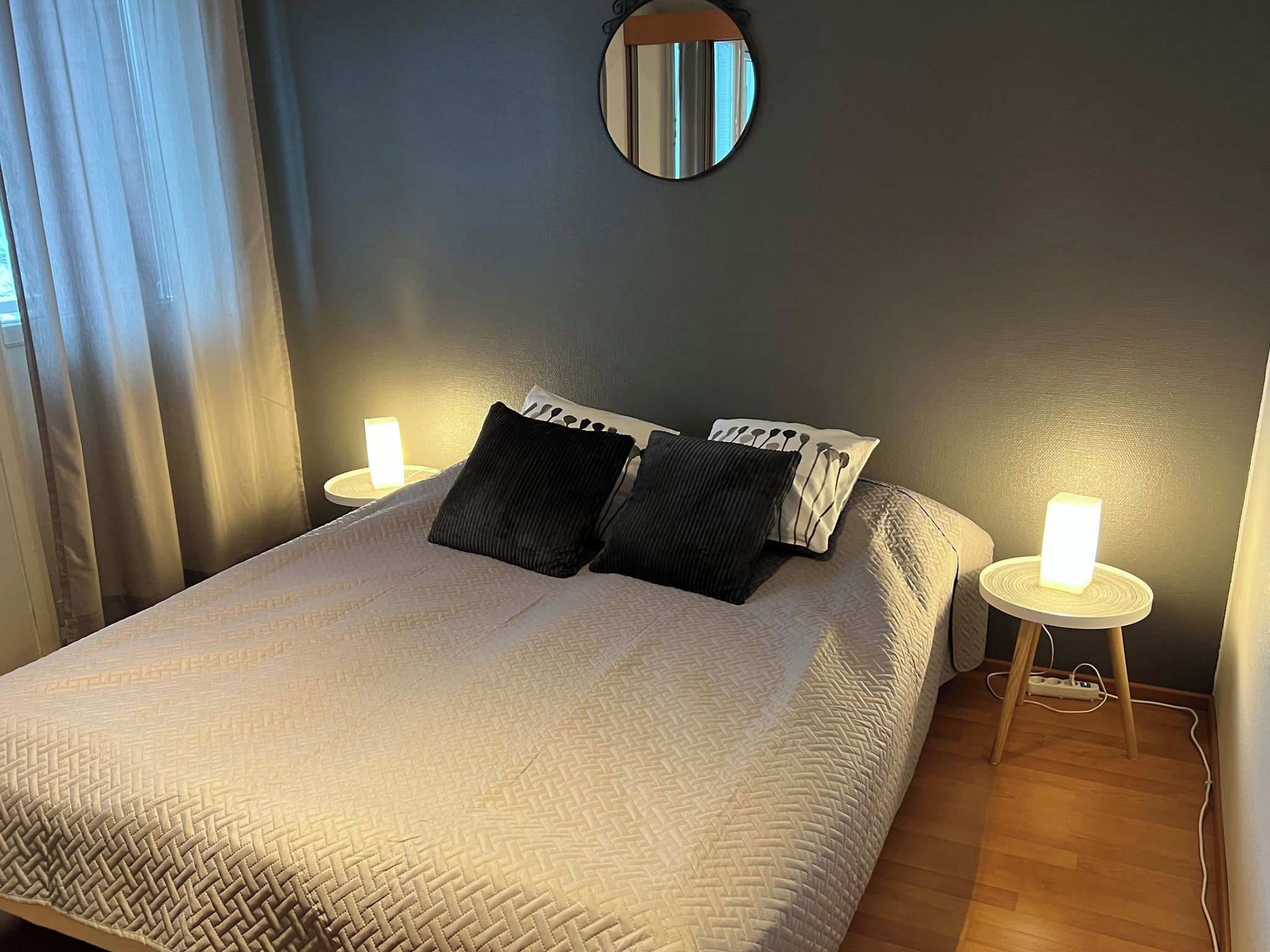 A neatly made bed with a white textured bedspread, two black pillows, and a round mirror on the wall. There are two bedside tables with white lamps.