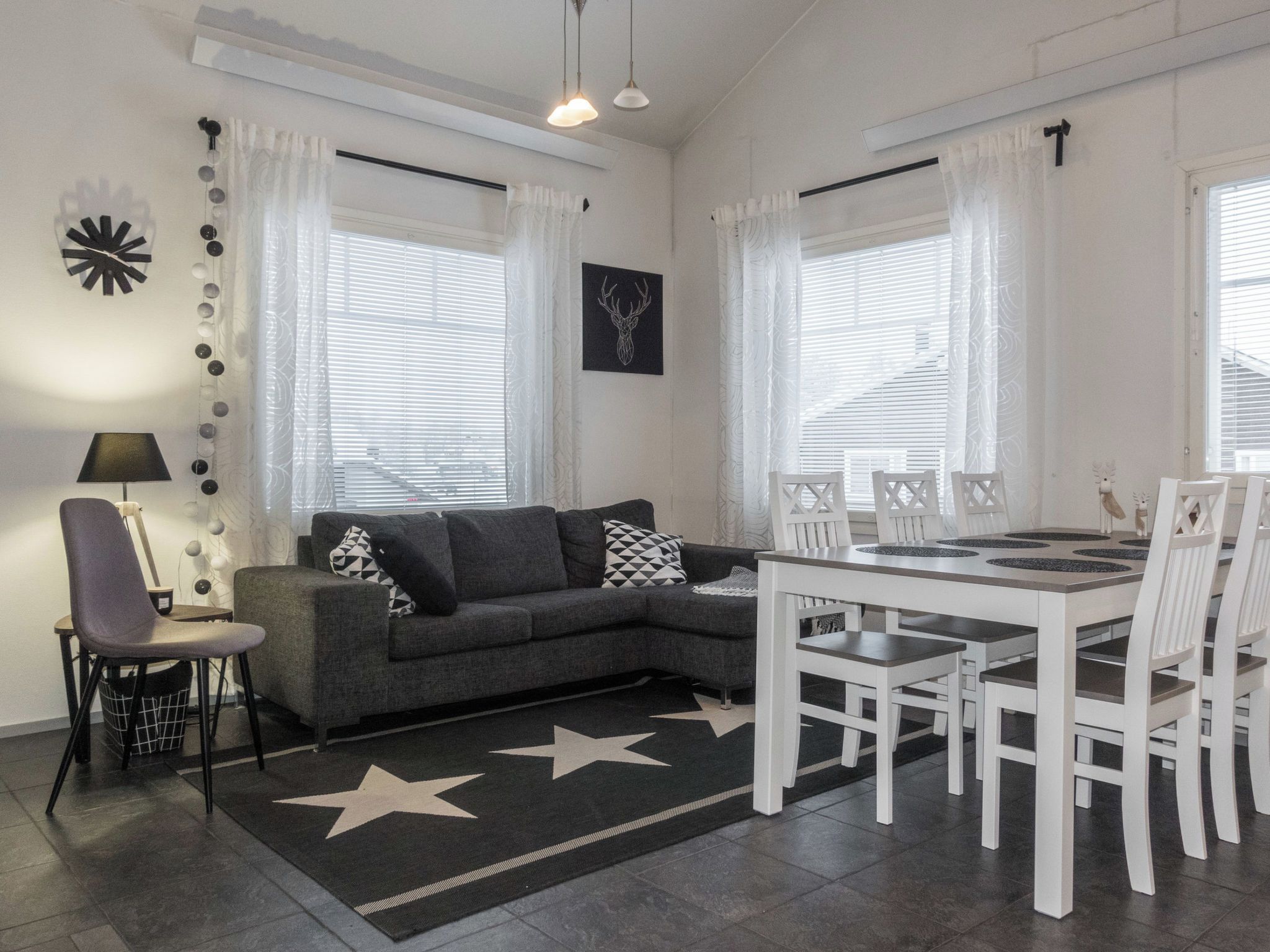 En moderne stue med en grå sofa, et hvidt spisebord med stole, et tæppe med stjernemønster og et vægophængt ur.