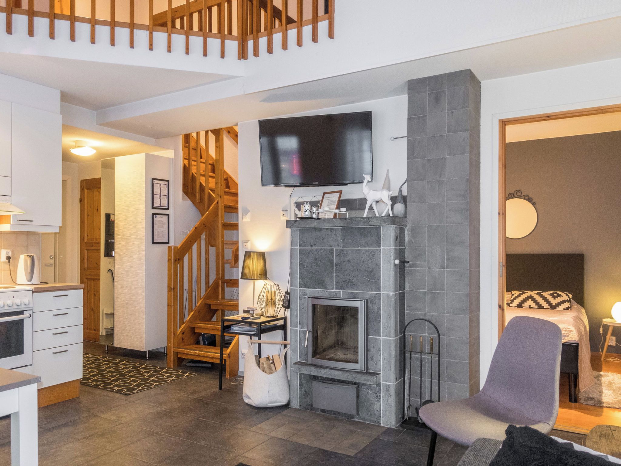 A modern living room with a fireplace, a flat-screen TV mounted above it, a dining area with a kitchen in the background, and a staircase leading to another level.