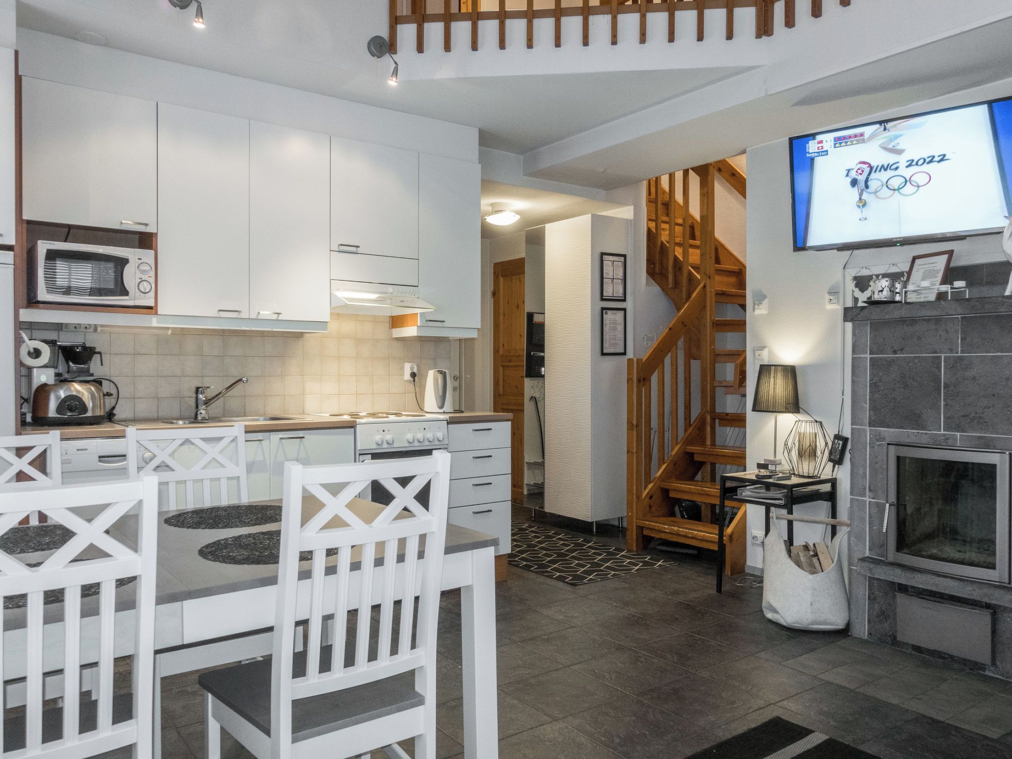 A modern kitchen with white cabinetry, a dining area with white chairs, a wooden staircase, and a television displaying the Tokyo 2022 Olympics logo.