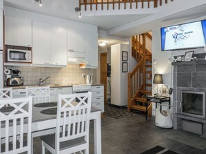 A modern kitchen with white cabinetry, a dining area with white chairs, a wooden staircase, and a television displaying the Tokyo 2022 Olympics logo. - Keskuskuja b4 by Interhome (Kittilä)