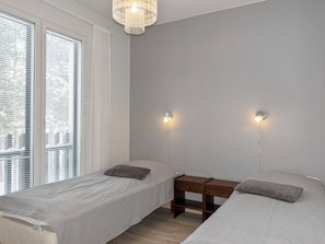 A bedroom with two single beds, a nightstand, a wall-mounted light fixture, and a window with blinds. - Golfstar 402 by Interhome (Haukipudas)