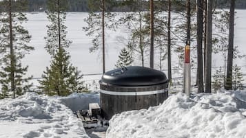 Lumipeitteinen ulkoalue, jossa on sylinterimäinen rakennelma, mahdollisesti sauna, ja sitä ympäröivät lumikinokset ja puut.