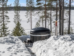 Área externa coberta por neve com uma estrutura cilíndrica, possivelmente uma sauna, rodeada por montes de neve e árvores.