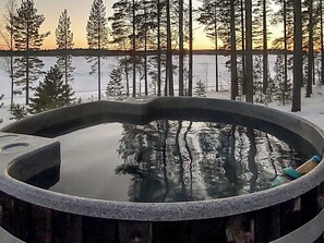 Een hottub omgeven door een met sneeuw bedekte grond en bomen, met een ondergaande zon op de achtergrond.