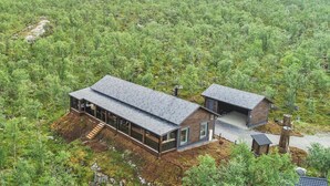 Een moderne cabin met een grote glazen gevel en een houten buitenkant, omgeven door dicht bos en een heuvel.