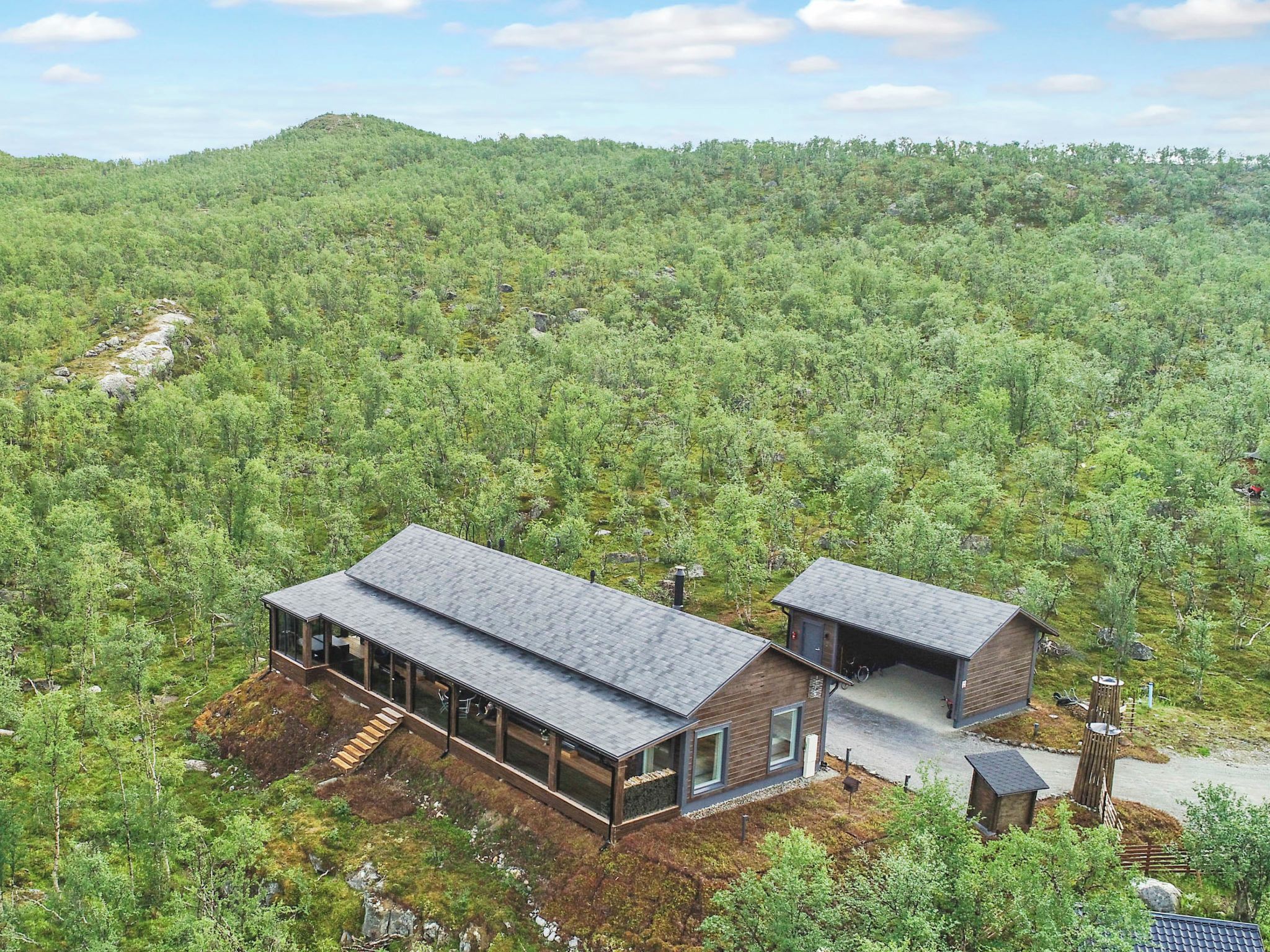Une cabane moderne dotée d’une grande terrasse et d’un garage, entourée d’une forêt dense.