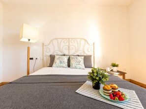 A neatly made bed with a grey bedspread, a white headboard, and two pillows. There is a small table with a plant and a plate of food.