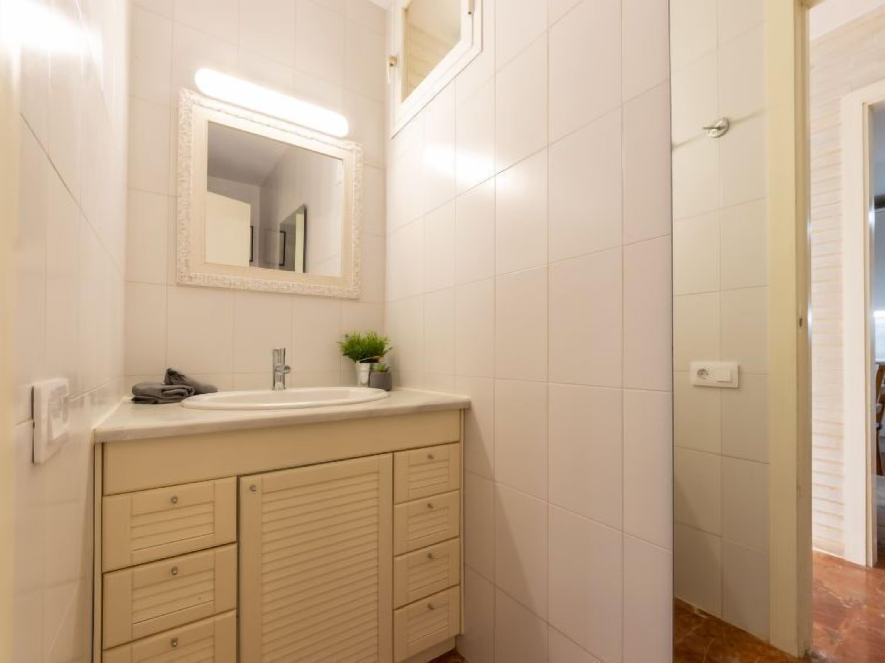 A bathroom with white tiled walls, a sink with a mirror above it, a small potted plant, and a door leading to another room.