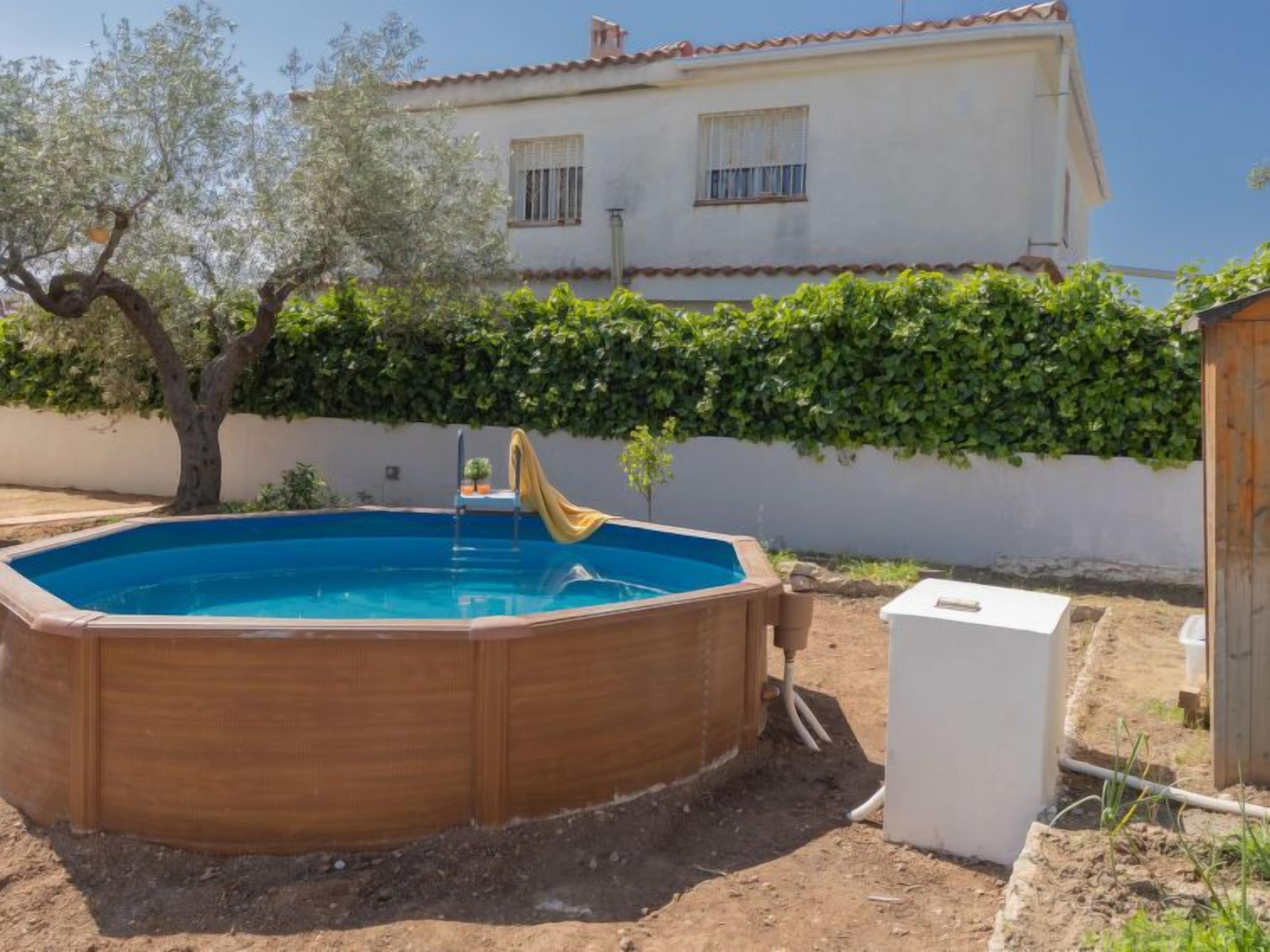 An outdoor swimming pool with a wooden deck, surrounded by a garden and a white house in the background.