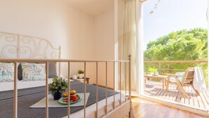 A balcony with a wooden deck, a bed with a grey mattress and floral pillows, and a view of greenery outside. - Mar Roig by Interhome (L'Ampolla)