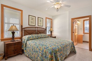 A bedroom with a bed, two bedside lamps, a wooden nightstand, a ceiling fan, and a door leading to a bathroom. - Soundbreeze Sunset: Golf and sound views, hot tub, dog friendly, elevator. (Nags Head)
