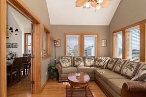 A living room with a sectional sofa, wooden flooring, and large windows with blinds.