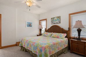 A bedroom with a bed, a nightstand, a lamp, a ceiling fan, and two wall-mounted paintings. - Soundbreeze Sunset: Golf and sound views, hot tub, dog friendly, elevator. (Nags Head)