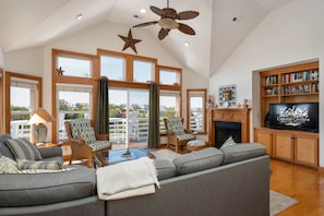A spacious living room with a large sofa, two armchairs, a coffee table, and a fireplace. There is a bookshelf and a television on the wall. - Soundbreeze Sunset: Golf and sound views, hot tub, dog friendly, elevator. (Nags Head)