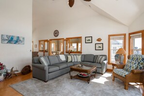 A spacious living room with a grey sectional sofa, a coffee table, and a wicker armchair. There are multiple windows with wooden frames and a large mirror on the wall. - Soundbreeze Sunset: Golf and sound views, hot tub, dog friendly, elevator. (Nags Head)