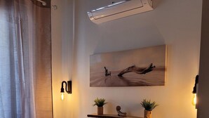 Un dormitorio con una cama, dos almohadas, una mesita de noche, un estante en la pared con plantas, un cuadro y un equipo de aire acondicionado.