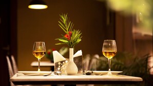 A dining table with two glasses of white wine, a vase with green foliage and a red flower, and a white napkin holder.