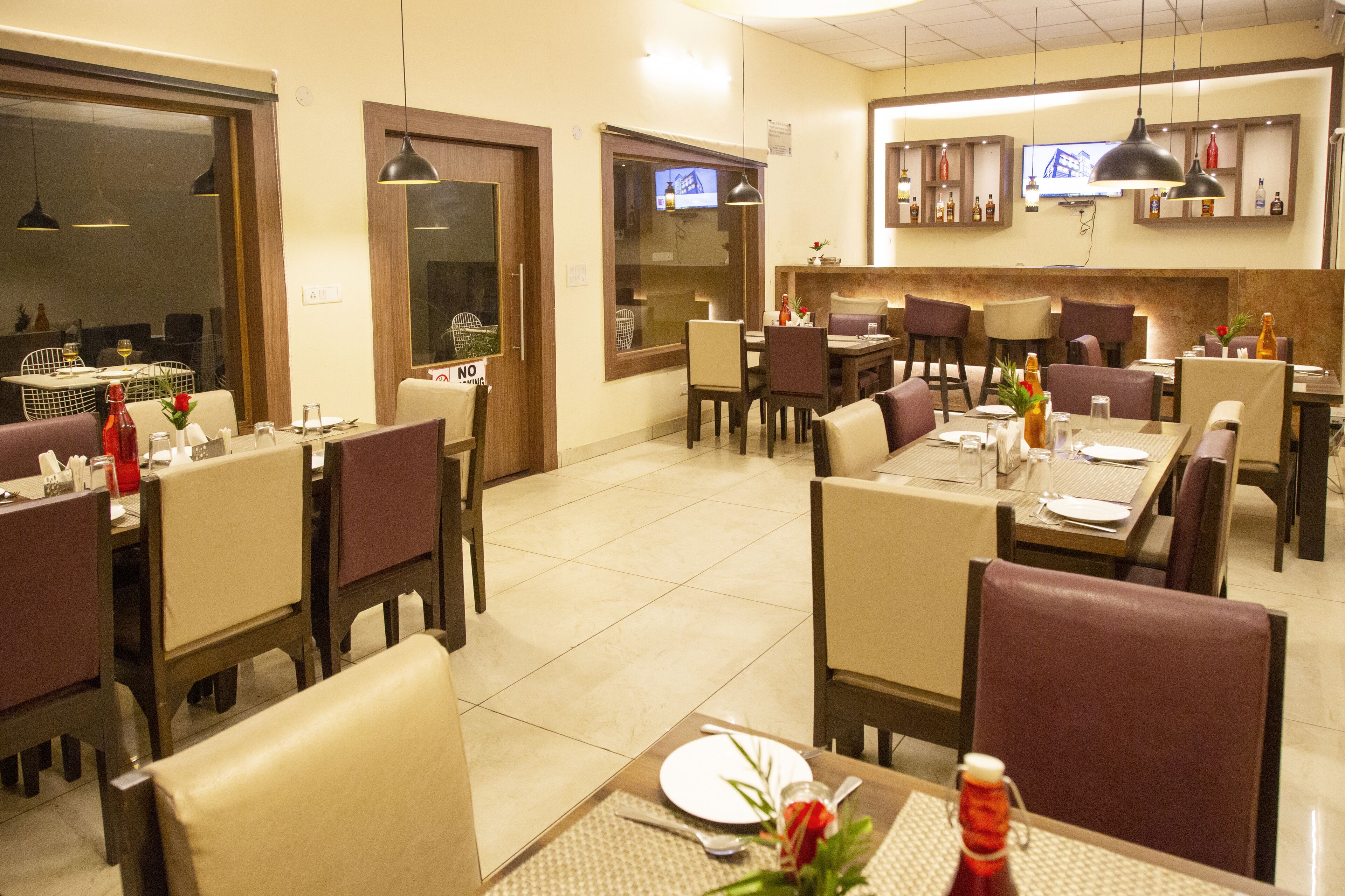 A restaurant with multiple dining tables and chairs, a television mounted on the wall, and a variety of bottles on shelves.
