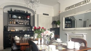 A dining area with a wooden table set for four, surrounded by chairs. A chandelier hangs from the ceiling, and a bookshelf filled with various items is visible in the background. - The Wadham (Weymouth)