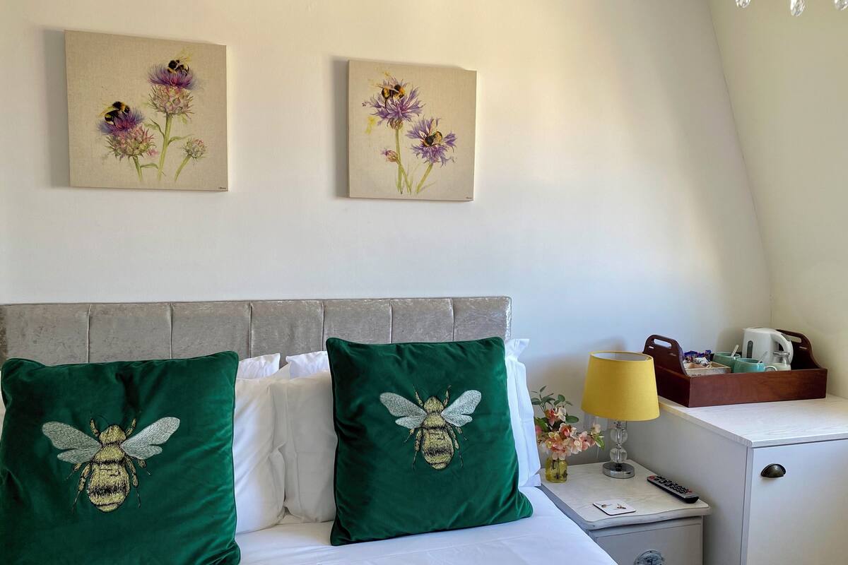 A hotel room with a bed, two green pillows with bee embroidery, a nightstand, a lamp, and a small cabinet.
