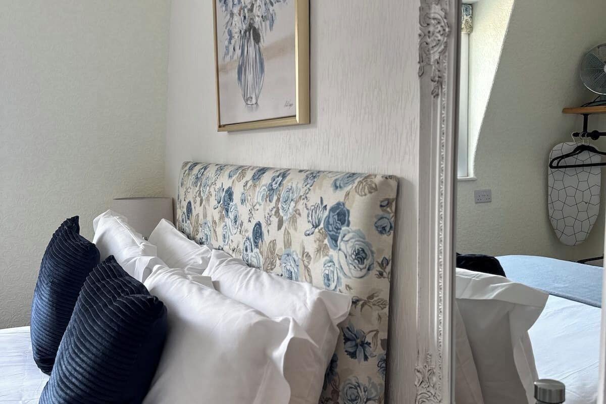 A neatly made bed with white and dark pillows, a floral-patterned headboard, a bedside table with a water bottle, and a framed artwork on the wall.