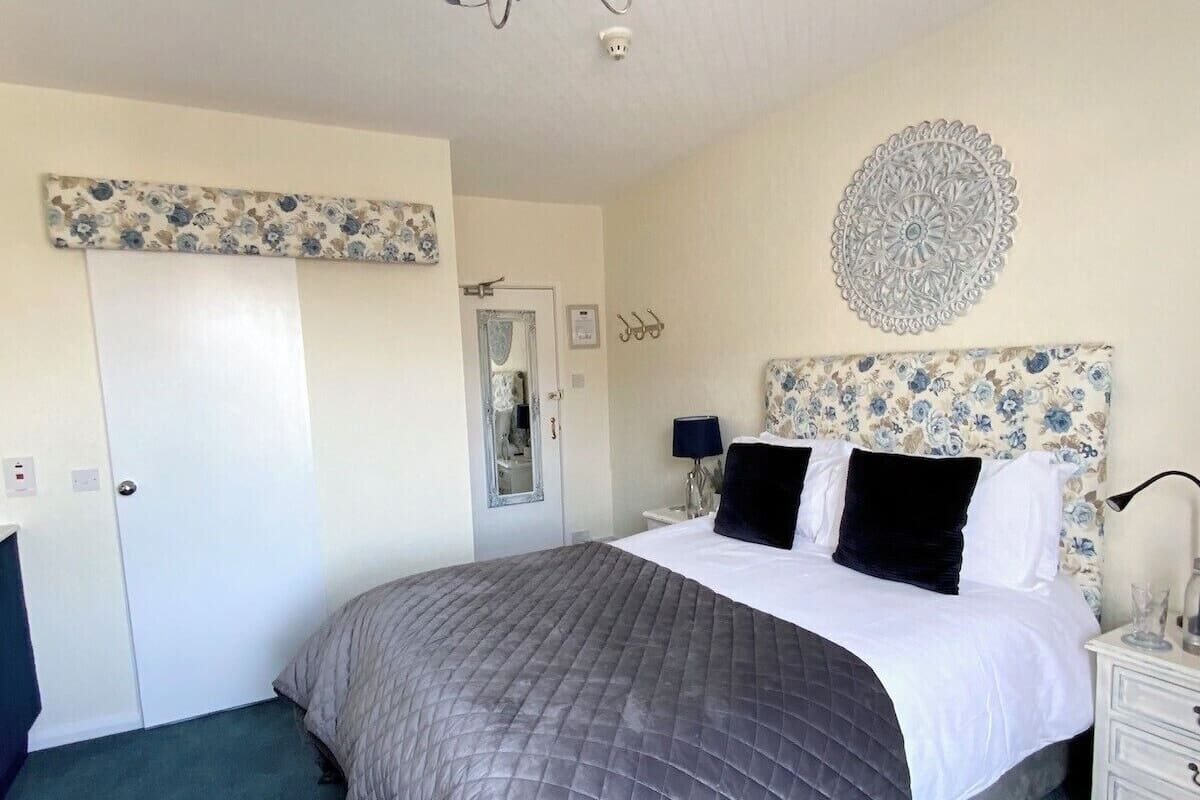 A bedroom with a large bed, a chandelier, a floral-patterned headboard, a mirror, and a nightstand with a lamp.