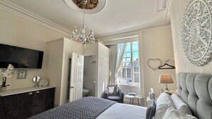 A hotel room with a large bed, a television mounted on the wall, a chandelier, and a decorative wall piece. - The Wadham (Weymouth)
