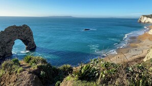 A coastal landscape with a prominent rock arch, sandy beach, and clear blue sea. - The Wadham (Weymouth)