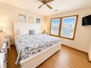A bedroom with a bed, a ceiling fan, a window with blinds, and a flat-screen TV mounted on the wall.