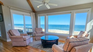 Een ruime woonkamer met een groot raam met uitzicht op het strand, ingericht met gestreepe banken en een ronde salontafel.