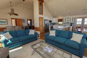 A spacious living room with two blue sofas, a glass coffee table, and a kitchen in the background. - A SHORE DELIGHT - ⛱️ Oceanside 5 BR home w/Private Pool, Hot Tub, and Pool Table ⛱️ (Corolla)