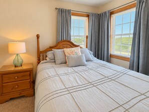 A bedroom with a wooden bed, a nightstand with a lamp, and two windows with curtains. - A SHORE DELIGHT - ⛱️ Oceanside 5 BR home w/Private Pool, Hot Tub, and Pool Table ⛱️ (Corolla)